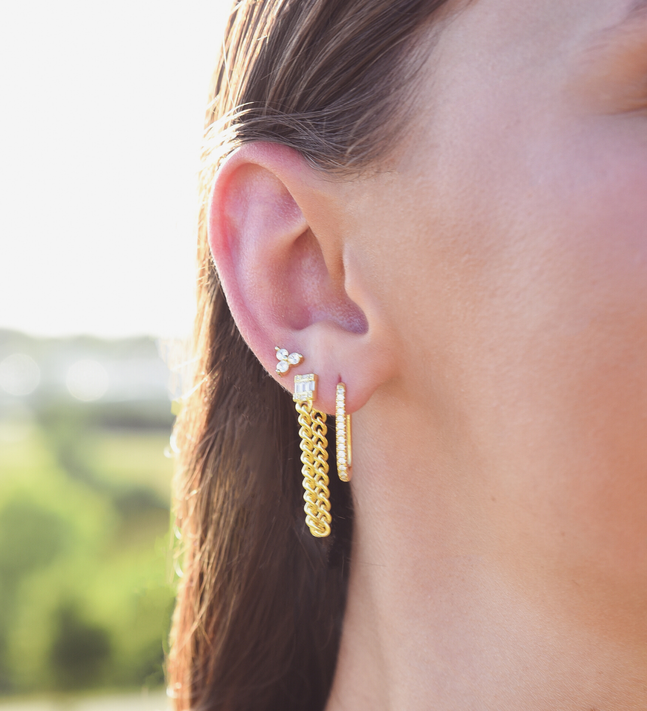 Side of Burdlife Model's head displaying 3 earrings including the Luxe Oval Hoops, the Refined Single Chain Earring and the Modish Triple Crystal Studs. 