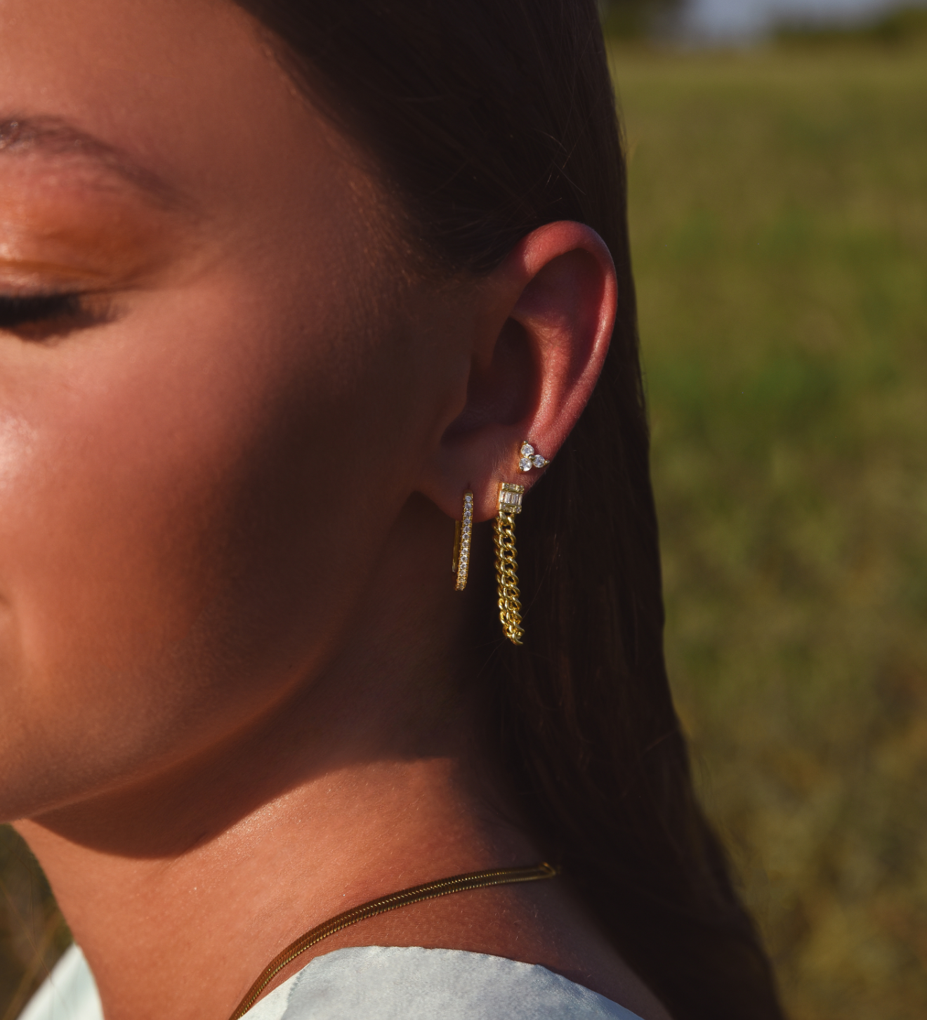 Side of Burdlife Model's head displaying 3 earrings including the Luxe Oval Hoops, the Refined Single Chain Earring and the Modish Triple Crystal Studs. 